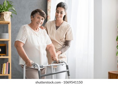 Happy Pensioner Trying To Walk In A Senior Home With Support Of The Nurse