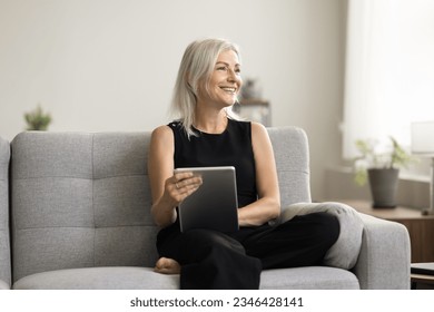 Happy pensive blonde entrepreneur woman sitting on sofa, holding tablet computer for online connection, looking away, smiling, enjoying work from home, thinking on freelance work project - Shutterstock ID 2346428141