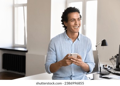 Happy pensive handsome young business leader man holding cellphone, using online app for Internet communication in office, chatting, standing at work table, looking away, thinking, dreaming, smiling - Powered by Shutterstock