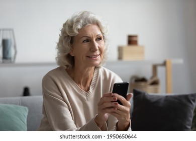 Happy Pensive Mature Lady Holding Smartphone At Home, Looking Away And Smiling. Elderly 60s Woman Thinking Over Phone Video Call For Talk To Doctor, Using Online Telemedicine App On Cell, Chatting