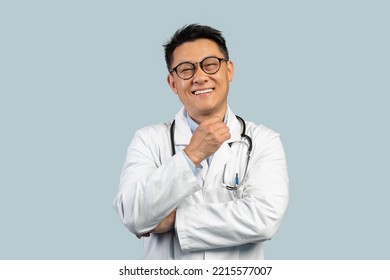 Happy Pensive Middle Aged Chinese Male Doctor In White Coat, Glasses Look At Camera, Isolated On Blue Background. Diagnostics, Ad And Offer, Treatment, Health Care And Medicine Due Covid-19 Pandemic