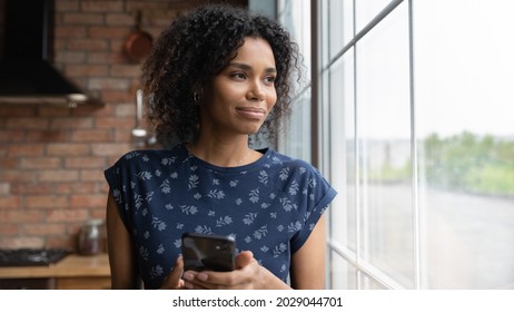 Happy Pensive Young African American Woman With Smartphone Looking Away Out Of Window, Smiling, Holding Mobile Phone, Thinking Of Good News From Text Message, Online Chat, Telephone Call