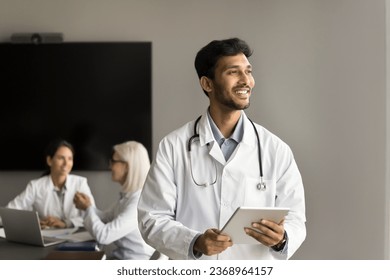 Happy pensive young Indian doctor standing for camera with meeting colleagues in background, using tablet computer, looking away with toothy smile, positive thoughts, successful medical career - Powered by Shutterstock