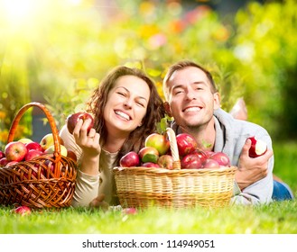 Happy People Eating Organic Apples In Autumn Garden.Healthy Food.Outdoors.Park. Basket Of Apples.Harvest Concept .Smiling Couple Relaxing On Grass