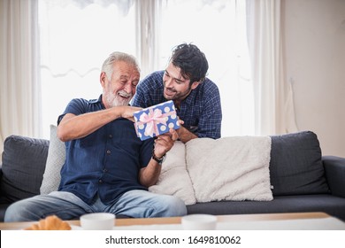 Happy people family concept - Old senior old man opens the gift box the son gave. Young man hugs his elderly father. Two men sitting on the sofa in the house. - Shutterstock ID 1649810062