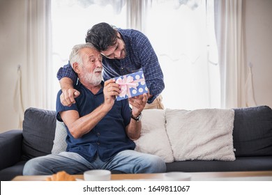 Happy people family concept - Old senior old man opens the gift box the son gave. Young man hugs his elderly father. Two men sitting on the sofa in the house. - Powered by Shutterstock