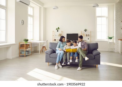 Happy people sitting together on sofa in modern interior of new house or studio apartment. Family with kids reading book on comfy gray couch in the living-room and enjoying quiet leisure time at home