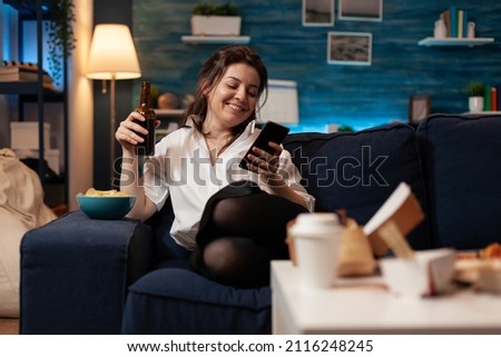 Happy person having fun browsing smartphone while drinking beer sitting confortable on couch after tasty burger takeaway fast food dinner. Smiling woman browsing social media in living room.