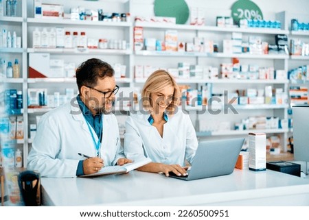Happy pharmacist using laptop while her colleagues is taking notes in a pharmacy.