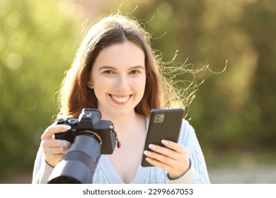 Happy photographer looks at you holding cell phone and mirrorless camera in a park