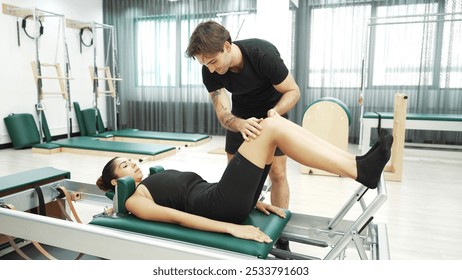 Happy pilates instructor assisting a client in performing a leg press exercise on a reformer for improving core stability. Sporty girl using pilates machine posing arm circle on arc barrel. Habituate. - Powered by Shutterstock