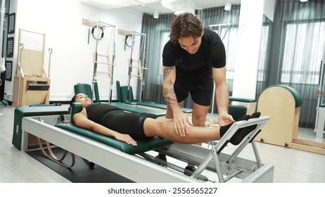 Happy pilates instructor assisting a client in performing a leg press exercise on a reformer for improving core stability. Sporty girl using pilates machine posing arm circle on arc barrel. Habituate. - Powered by Shutterstock