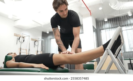 Happy pilates instructor assisting a client in performing a leg press exercise on a reformer for improving core stability. Sporty girl using pilates machine posing arm circle on arc barrel. Habituate. - Powered by Shutterstock