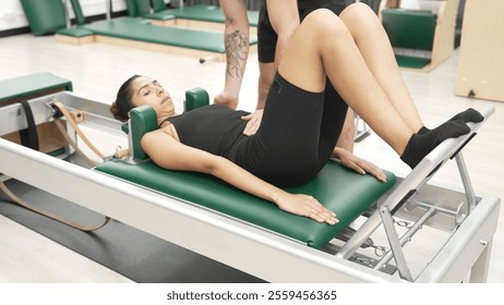 Happy pilates instructor assisting a client in performing a leg press exercise on a reformer for improving core stability. Sporty girl using pilates machine posing arm circle on arc barrel. Habituate. - Powered by Shutterstock