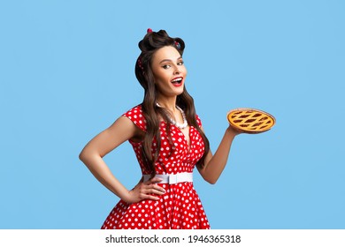 Happy Pin Up Woman In Polka Dot Red Dress Holding Homemade Sweet Pie On Blue Studio Background. Cheerful Young Housewife In Retro Style Dress With Yummy Pastry Dessert