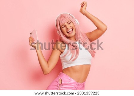 Happy pink-haired girl with headphones holds phone in one hand danciing while listening music, posing on pink background, copy space, delight concept 
