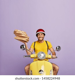 Happy piza delivery man busy with his duties, holds pile of carton boxes, drives scooter, wears helmet and yellow t shirt, isolated on purple background, works in pizzeria, represents restaurant well
