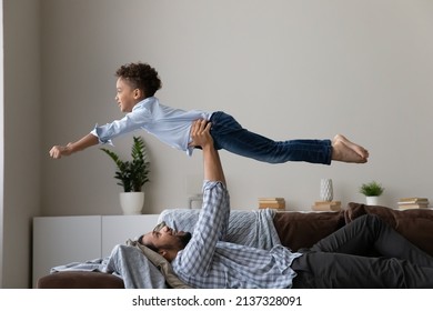 Happy playful African daddy playing sportive games with active little son, holding boy making airplane, super hero, doing acroyoga base, lifting kid up in air, laughing, resting on sofa - Powered by Shutterstock