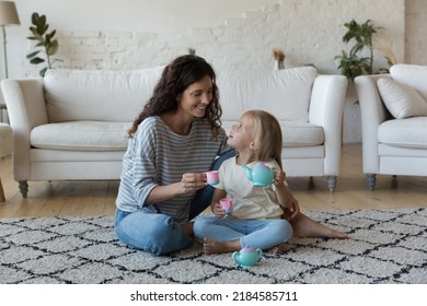 Happy playful caring mother and sweet little daughter girl playing tea party with tiny toy cups, teapot on heating floor, pretending drinking coffee in game at home, talking, laughing - Shutterstock ID 2184585711