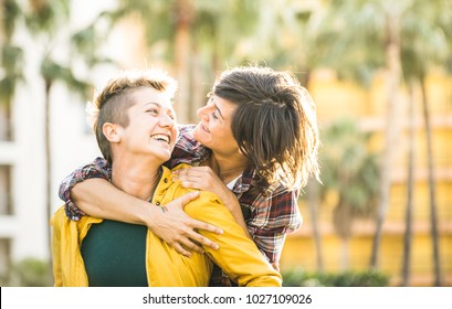 Happy playful girlfriends in love sharing time together at travel trip on piggyback hug - Women friendship concept with girls couple having fun on fashion clothes outdoors - Bright warm sunset filter