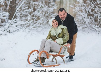 Happy Playful Mature Family Couple Sledding In Winter Park, Laughing And Having Fun Together, Positive Middle-aged Woman Sitting On Sled While Her Husband Pushing Her From Behind. Outdoor Activities