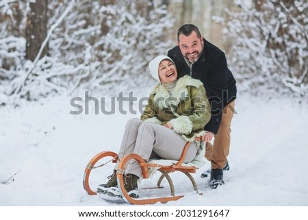 Happy playful mature family couple sledding in winter park, laughing and having fun together, positive middle-aged woman sitting on sled while her husband pushing her from behind. Outdoor activities