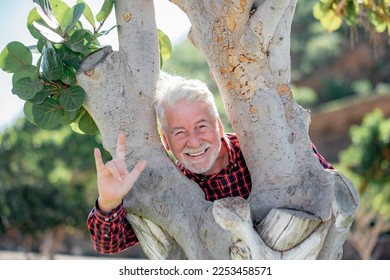 Happy playful senior bearded man partially hidden by a tree trunk in the park looking at camera smiling. Elderly grandfather enjoying free time and healthy retirement in outdoor