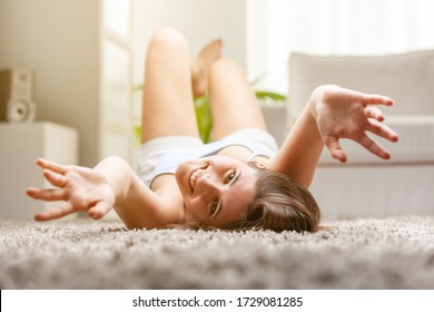 Happy Playful Young Woman Reaching To The Camera With Her Outstretched Hands As She Lies On Her Back On A Carpet In The Living Room In A Low Angle View