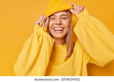 Happy playful young woman smiles broadly shows white teeth keeps hands on hat dressed in casual jumper expresses sincere emotions being in good mood isolated over yellow background. Joy concept - Shutterstock ID 2078634502