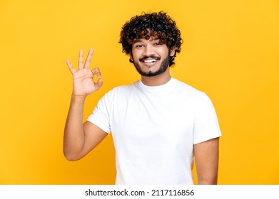 Happy Pleasant Indian Or Arabian Curly-haired Guy In White T-shirt, Showing Ok Sign While Standing On Isolated Orange Background, Looks At Camera, Smiling Friendly