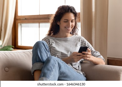 Happy pleasant millennial woman relaxing on comfortable couch, holding smartphone in hands. Smiling young lady chatting in social networks, watching funny videos, using mobile applications at home. - Shutterstock ID 1646346130