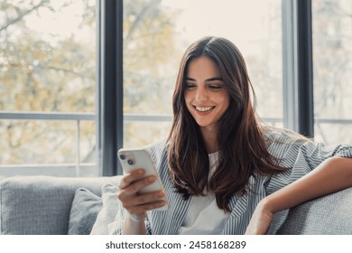 Happy pleasant millennial woman relaxing on comfortable couch, holding smartphone in hands. Smiling young lady chatting in social networks, watching funny videos, using mobile applications at home. - Powered by Shutterstock