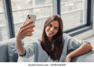 Happy pleasant millennial woman relaxing on comfortable couch, holding smartphone in hands. Smiling young lady taking a selfie, posting on social media, using mobile applications at home. - Powered by Shutterstock