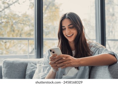 Happy pleasant millennial woman relaxing on comfortable couch, holding smartphone in hands. Smiling young lady chatting in social networks, watching funny videos, using mobile applications at home. - Powered by Shutterstock