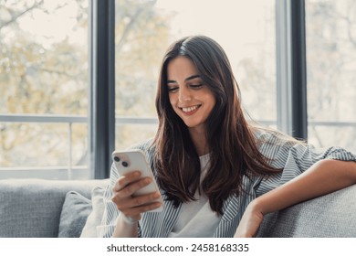 Happy pleasant millennial woman relaxing on comfortable couch, holding smartphone in hands. Smiling young lady chatting in social networks, watching funny videos, using mobile applications at home. - Powered by Shutterstock