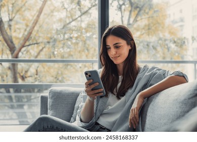 Happy pleasant millennial woman relaxing on comfortable couch, holding smartphone in hands. Smiling young lady chatting in social networks, watching funny videos, using mobile applications at home. - Powered by Shutterstock