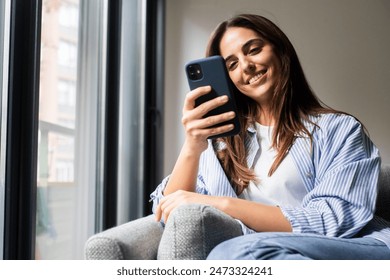 Happy pleasant millennial woman relaxing on comfortable couch, holding smartphone in hands. Smiling young lady chatting in social networks, watching funny videos, using mobile applications at home. - Powered by Shutterstock