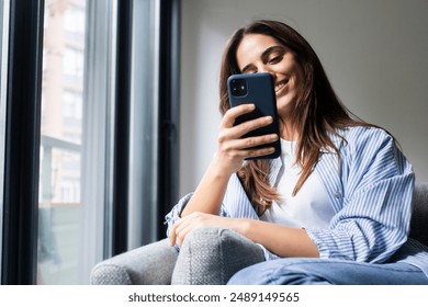 Happy pleasant millennial woman relaxing on comfortable couch, holding smartphone in hands. Smiling young lady chatting in social networks, watching funny videos, using mobile applications at home. - Powered by Shutterstock