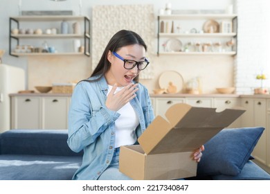 Happy And Pleasantly Surprised Young Beautiful Asian Woman Sitting On Sofa At Home And Opening Cardboard Box, Gift. I Received An Unexpected Package In The Mail.