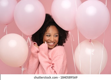 Happy pleased biracial woman with curly hair closed eyes, holding a lot of balloons, enjoys cool party, wears pink sweater, celebrates birthday, standing over studio pink background. Festive event