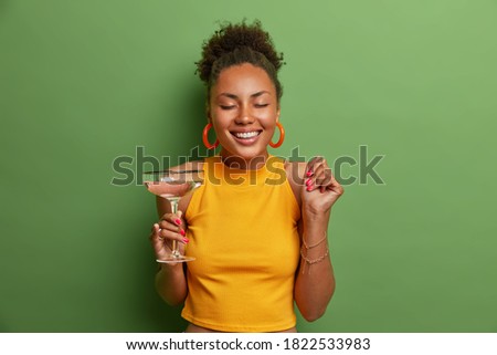 Happy pleased dark skinned woman closes eyes and smiles toothily raises clenched fist and drinks cold cocktail has happy mood dressed in casual outfit poses in studio against green background