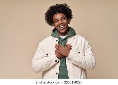 Happy pleased excited grateful African American teen guy holding hands on chest expressing gratitude, feeling trust hope love in heart, impressed with kindness isolated on beige background.