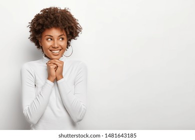 Happy pleased woman keeps hands together under chin, looks aside with joy, enjoys funny time, recalls nice memories, has toothy smile, wears hoop silver earrings and white jumper. Blank space right