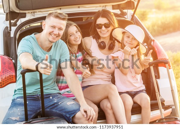 Happy pleasured family sitting on a trunk\
of a car with suitcases on a travel\
vacation