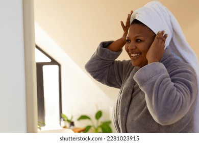 Happy plus size african american woman wearing robe looking at herself in bathroom mirror. Self care, health and beauty, body inclusivity and lifestyle, unaltered. - Powered by Shutterstock