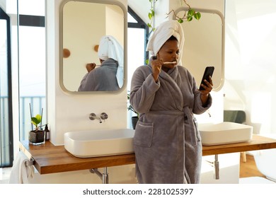 Happy plus size african american woman wearing robe, brushing teeth and using smartphone in bathroom. Self care, health and beauty, body inclusivity, communication and lifestyle, unaltered. - Powered by Shutterstock