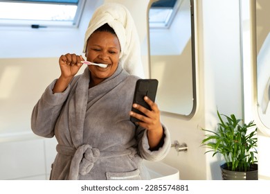 Happy plus size african american woman wearing robe, brushing teeth and using smartphone in bathroom. Self care, health and beauty, body inclusivity, communication and lifestyle, unaltered. - Powered by Shutterstock