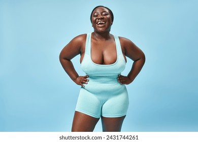 happy plus size african american woman in blue sportswear with hands on hips on matching backdrop - Powered by Shutterstock