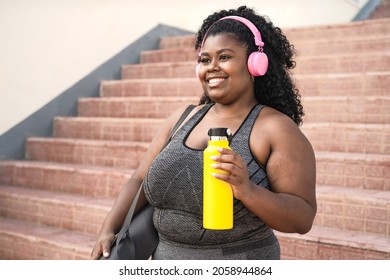 Happy plus size African woman doing workout routine while listening music with wireless headphones outdoor - Powered by Shutterstock