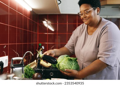 Happy plus size female of 60s with dark skin washing vegetables in sink with tap water, holding fresh aubergine and cabbage, cooking healthy dinner, enjoying process and harvest from her garden - Powered by Shutterstock